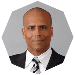 A man in a suit and tie poses for a formal portrait against a plain gray background. The image is cropped into a polygonal shape.