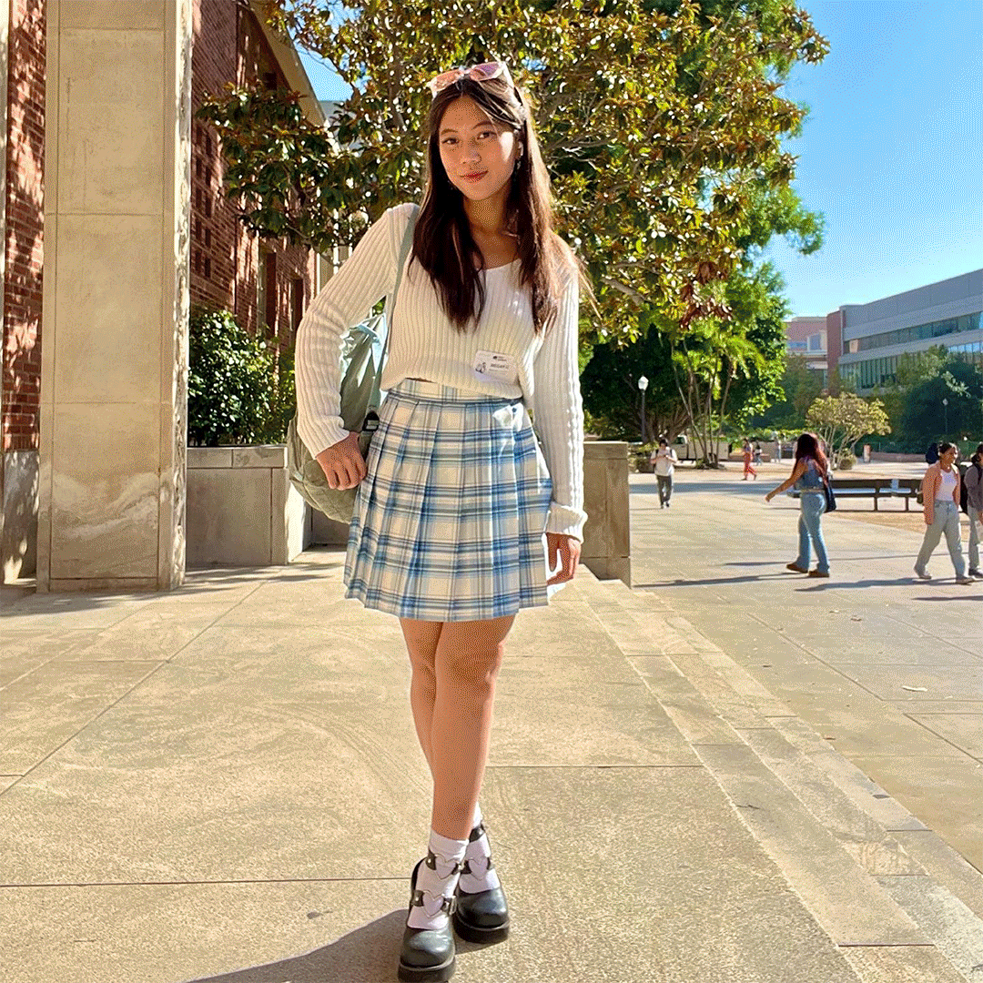 A young woman stands outdoors in sunlight at UCLA College, wearing a white ribbed sweater, a blue plaid skirt, and chunky white boots. Trees, a brick building, and a few people walking are visible in the background.