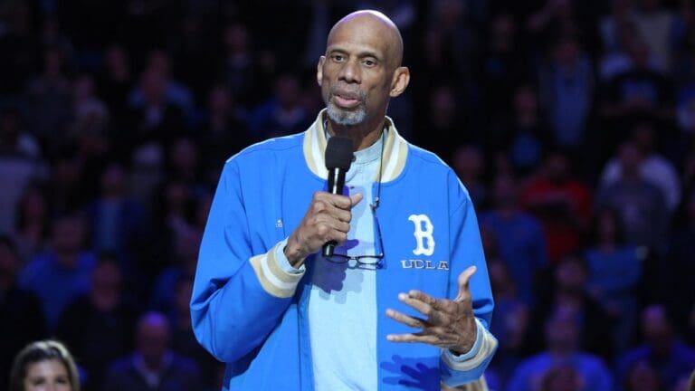 A tall man wearing a blue UCLA jacket holds a microphone and speaks to an audience. He stands in front of a blurred crowd, gesturing with his hand, with glasses hanging from his neck.