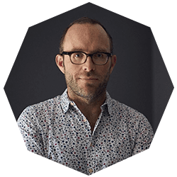 A man with short hair and glasses wearing a patterned button-up shirt poses against a dark background, framed by a geometric octagonal border.