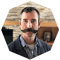 A man with short dark hair and a large, curled mustache is wearing a checked shirt and a blazer, posing in front of a stone wall and shelves.