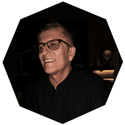 A person with short hair and glasses smiles while sitting indoors, wearing a dark shirt. The image has a dark background and is framed within an octagonal border.