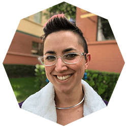 Smiling person with short dark hair styled upward and pink highlights, wearing clear glasses, a silver necklace, and a white-collared top, standing outdoors in front of a building and greenery.