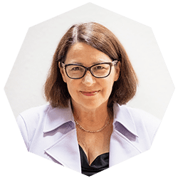 A woman with straight brown hair and glasses is smiling at the camera. She is wearing a light purple blazer, a black top, and a gold necklace, set against a plain, light background.