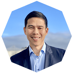 A man in a suit smiles at the camera against a blue sky background. The image is cropped in an octagonal shape.