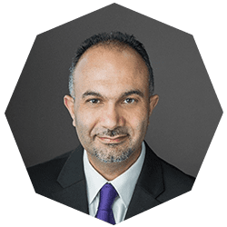 A man in a suit and purple tie smiles slightly in a professional portrait with a hexagonal frame and a gray background.
