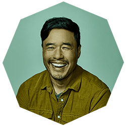 A man with short dark hair and a trimmed beard smiles widely while wearing a brown button-up shirt, set against a light green background with an octagonal border.