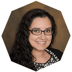 A woman with long, curly dark hair and glasses smiles at the camera. She is wearing a black top with a decorative, beaded neckline against a brown background.