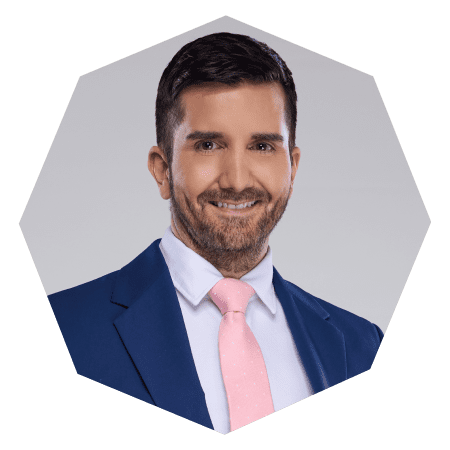 A man with short dark hair and a beard is wearing a blue suit, white shirt, and light pink tie, smiling at the camera against a light gray background in a hexagonal frame.