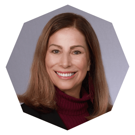 A woman with straight, shoulder-length brown hair and a maroon turtleneck sweater smiles at the camera against a gray background. The image is cropped in a hexagonal shape.
