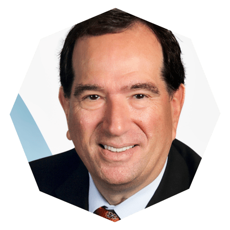 A middle-aged man with dark hair, wearing a dark suit, light blue shirt, and patterned tie, smiling at the camera against a light background.