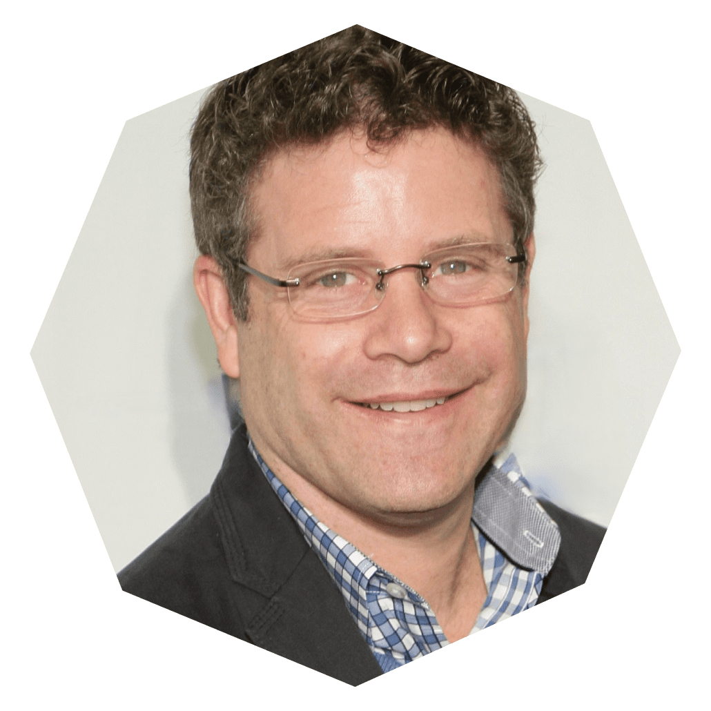 A man with short curly brown hair, glasses, and a checked shirt under a dark blazer smiles at the camera. The image is cropped into a hexagon shape with a plain light background.