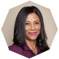 A woman with long straight black hair, wearing a purple blouse, smiles at the camera against a neutral background in a portrait photo with an octagonal frame.