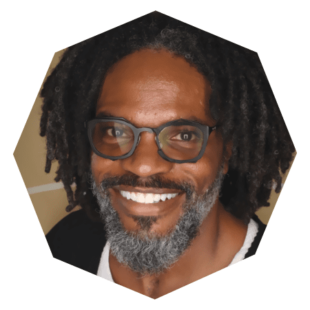 A smiling man with dark skin, glasses, gray and black beard, and shoulder-length curly hair, wearing a dark top, is photographed against a light background in a hexagonal frame.