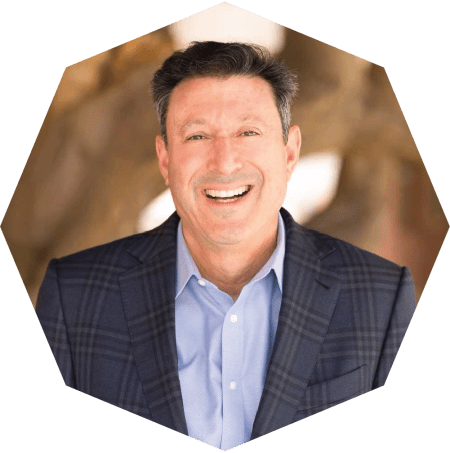 A man with short dark hair, wearing a blue plaid suit jacket and light blue shirt, smiles at the camera. The photo has a hexagonal border and a blurred neutral background.