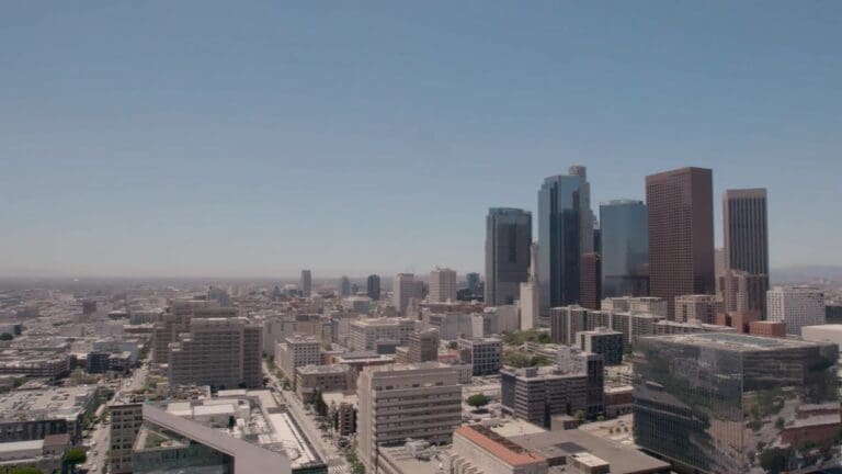 Aerial view of a city skyline with tall modern skyscrapers and various smaller buildings under a clear blue sky. The urban landscape stretches toward the horizon.