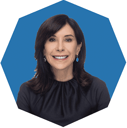 A woman with shoulder-length dark hair, wearing a dark top and earrings, smiles at the camera against a blue, octagonal background.