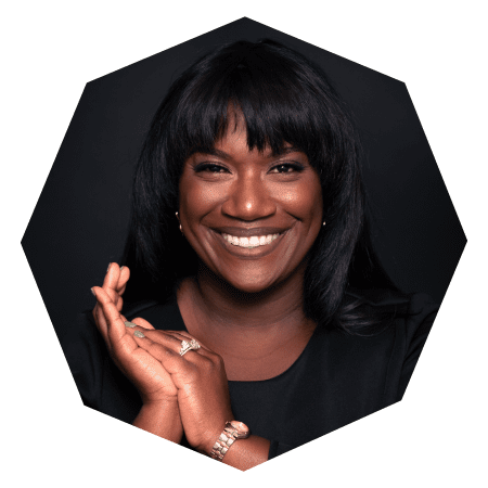 A woman with long black hair smiles warmly, wearing a black top, gold watch, and rings. She is clapping softly, posed against a dark background with a geometric octagonal crop.