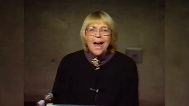 A person with short blonde hair, glasses, and a black top speaks enthusiastically at a podium with a microphone in front of a plain, grayish wall.
