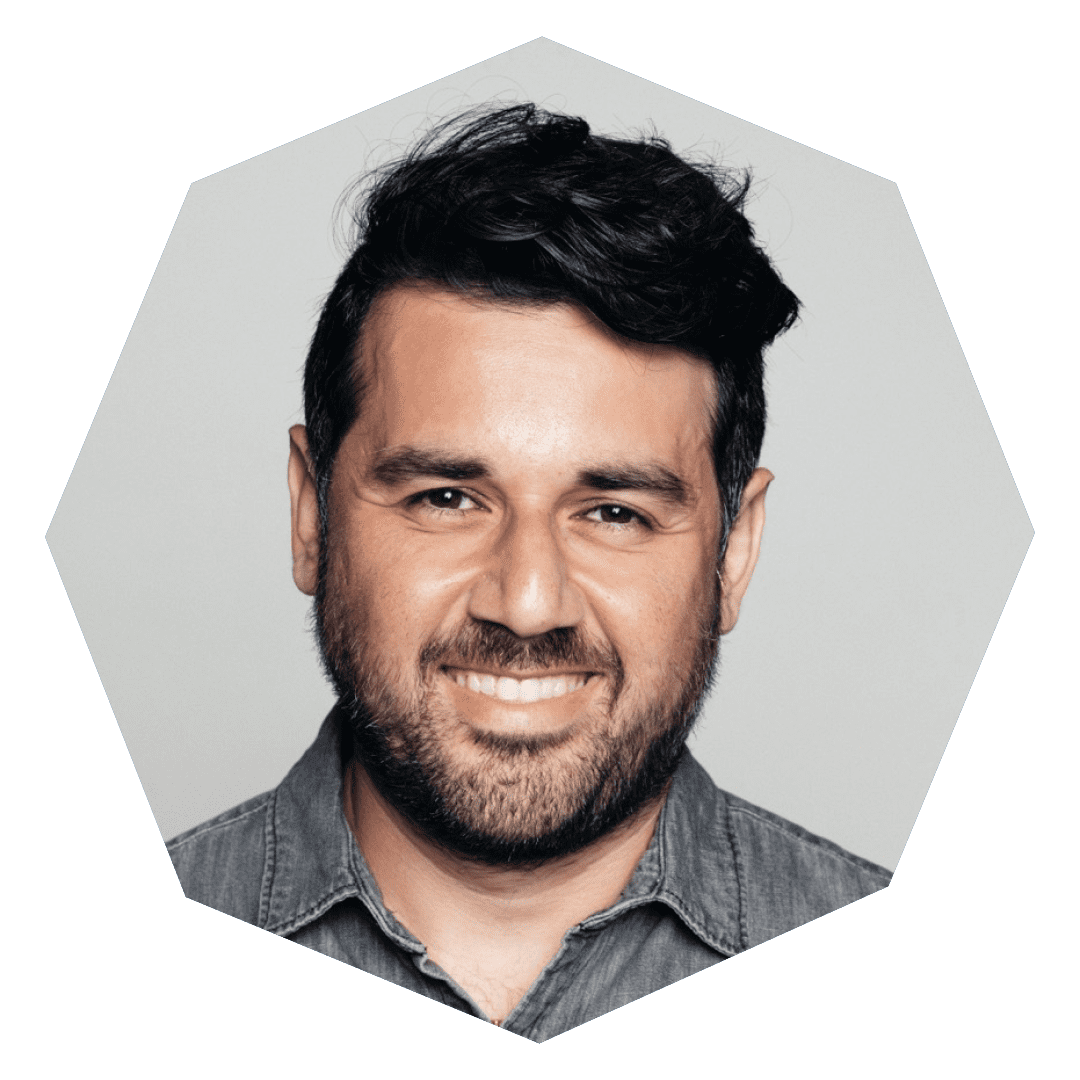 A man with dark hair and a beard smiles at the camera. He is wearing a gray button-up shirt and is pictured against a light gray background within an octagonal frame.