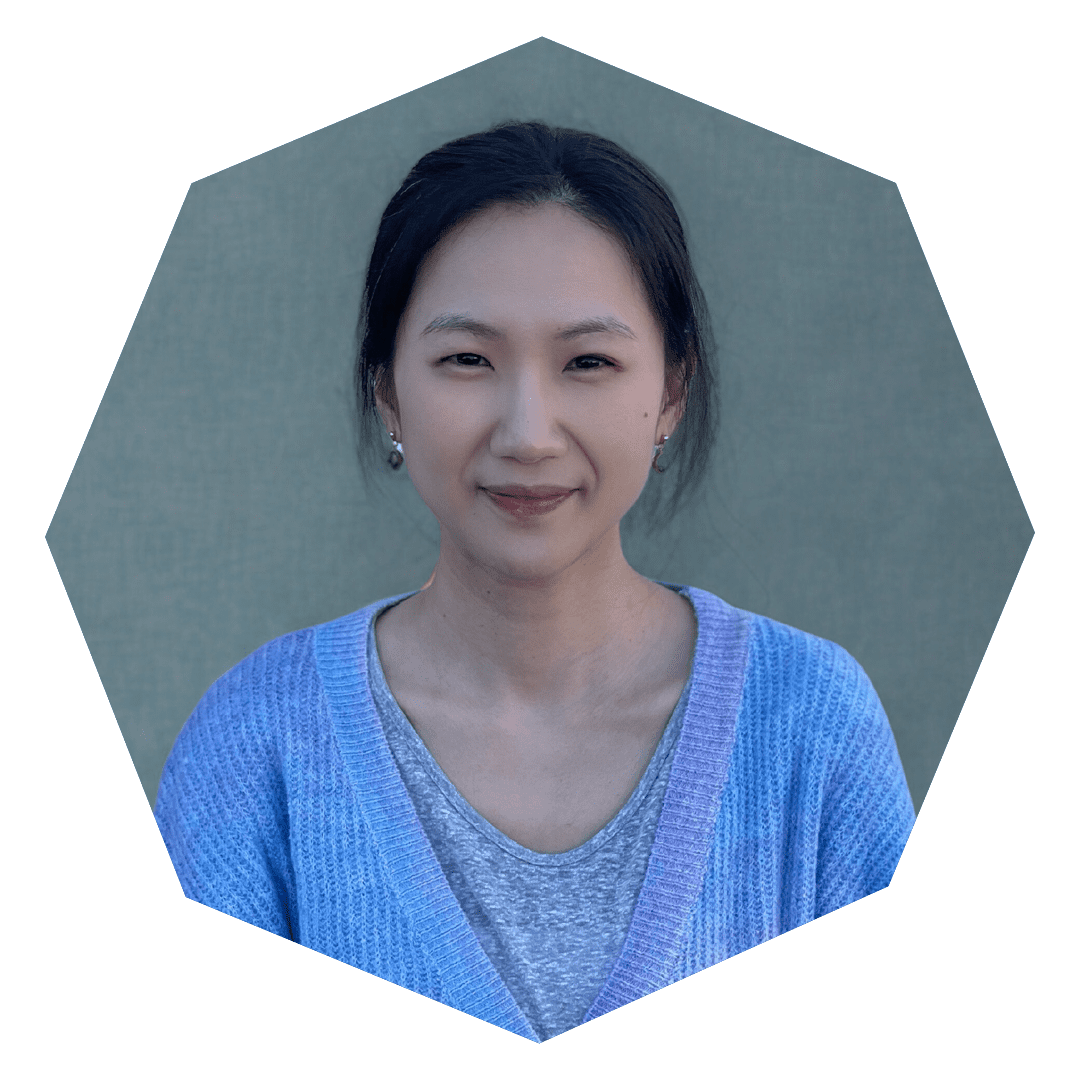 A person with long dark hair pulled back, wearing a light blue cardigan over a gray shirt, smiles softly against a muted green background. The image is cropped into an octagonal shape.