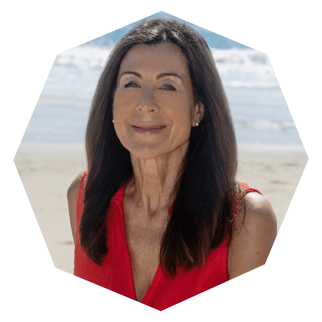 A woman with long brown hair wearing a sleeveless red top smiles while standing on a sandy beach with the ocean and waves in the background.