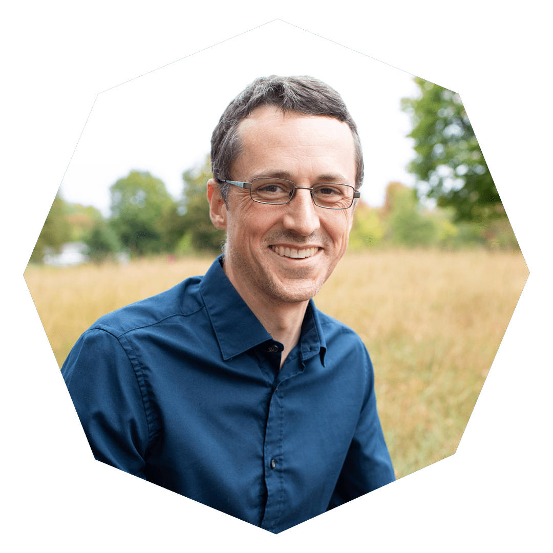 A man with short brown hair and glasses, wearing a dark blue button-up shirt, smiles while sitting outdoors in a grassy field with trees in the background. The image has a hexagon-shaped border.