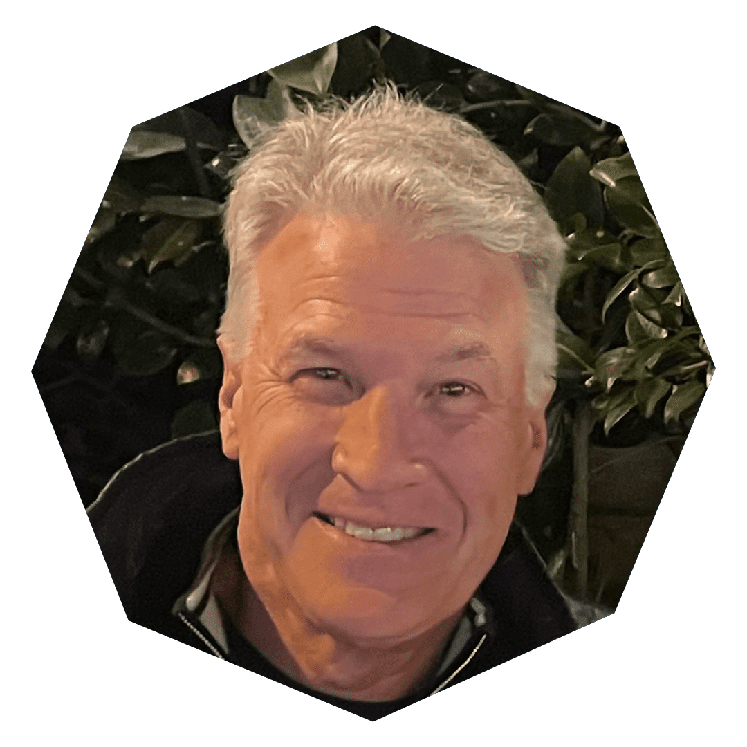 An older man with gray hair smiles at the camera, standing in front of dark green foliage. The image is cropped in a hexagonal shape.