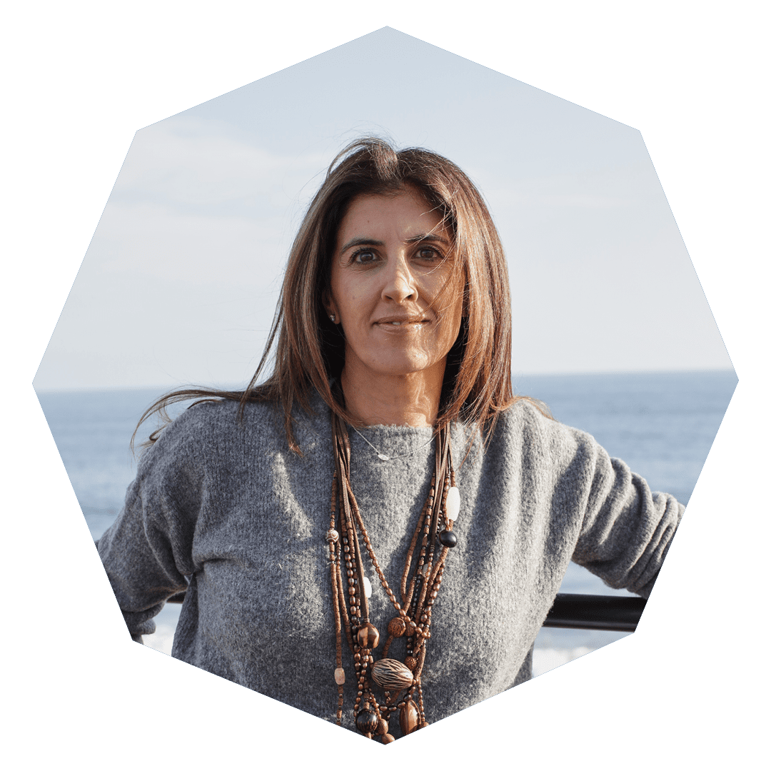 A woman with straight brown hair, wearing a gray sweater and layered beaded necklaces, stands outdoors with the ocean in the background. The image is framed in a hexagonal shape.