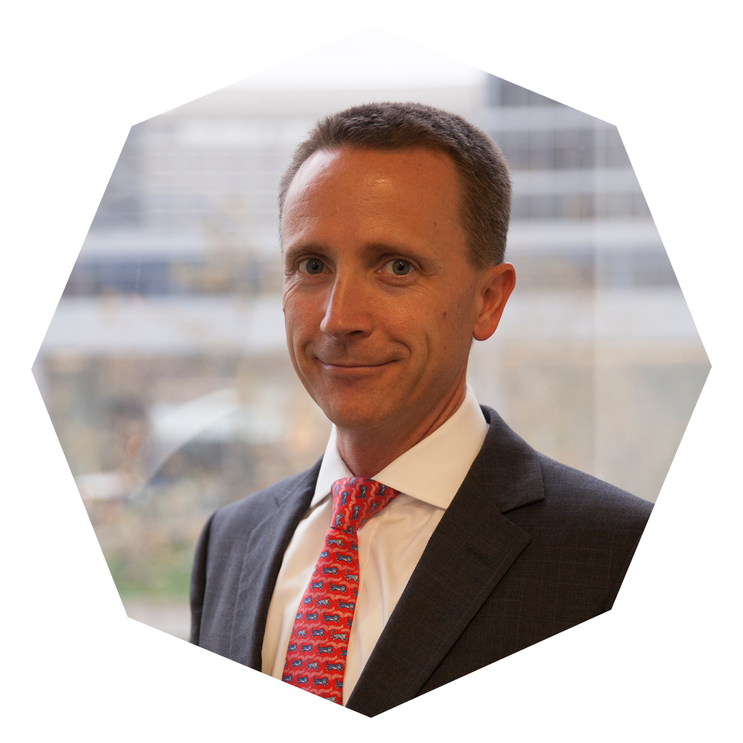 Aaron Bendikson in a blue suit, a white shirt and a red tie. The photo is framed in an eight-sided molecule.