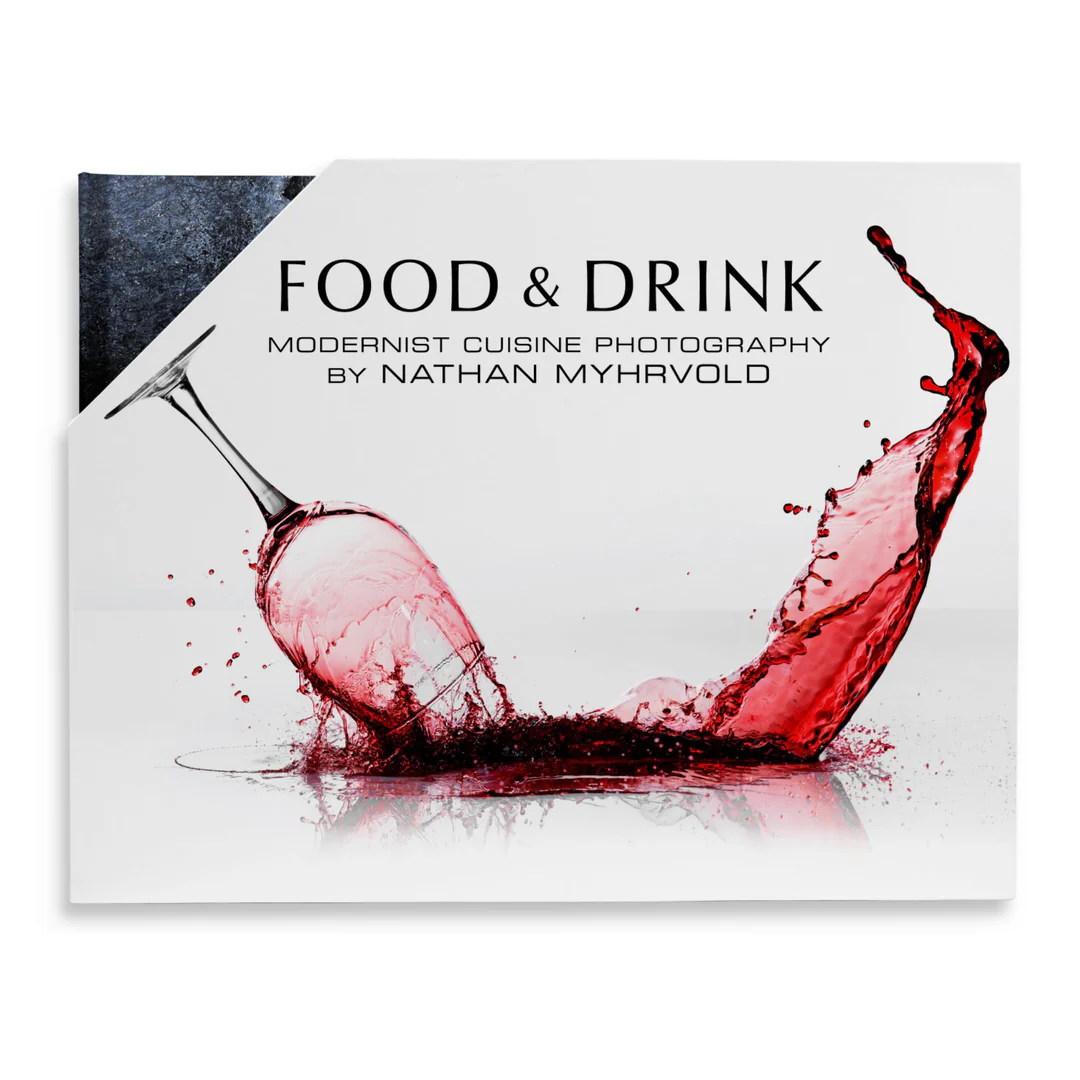 A wine glass tips over, spilling red wine with a dramatic splash. The text reads: FOOD & DRINK, Modernist Cuisine Photography by Nathan Myhrvold.