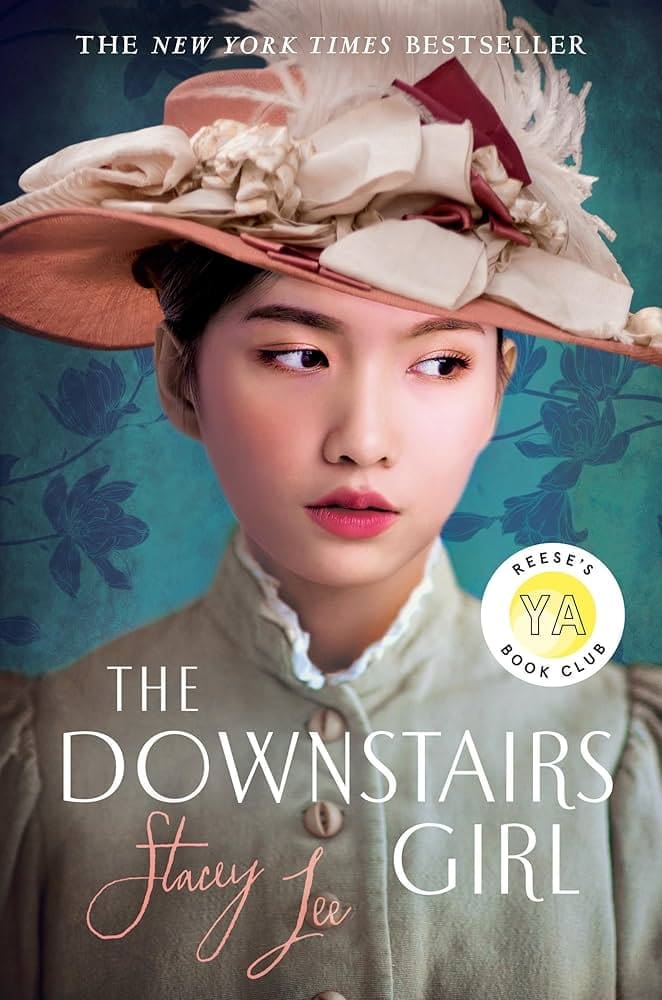 Book cover of The Downstairs Girl by Stacey Lee, featuring a young Asian woman wearing a vintage pink hat with flowers and a high-collared blouse against a teal floral background.