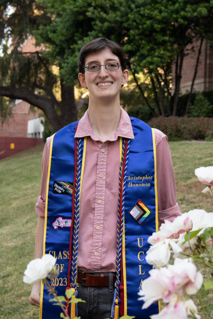 Christopher posing behind flowers on campus with his decorated graduation sash and cords. They represent OutWrite Newsmagazine, his disabled and queer identities, UCLA Student Media and the Disability Studies minor.